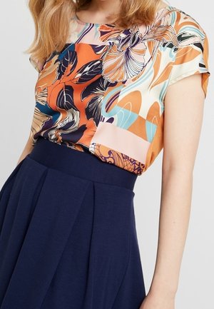 Vrouw draagt een meerkleurige abstracte kortemouwen top die in een hooggetailleerde donkerblauwe geplooide rok is gestopt.