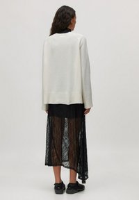 Lichtgrijze oversized gebreide trui met geribbelde zoom, gelaagd over een sheer zwarte kanten maxi-jurk, gecombineerd met zwarte sneakers.