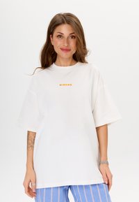 Witte oversized t-shirt met korte mouwen, met oranje "MIRONS" tekst op de voorkant. Zachte stof, ontspannen pasvorm, minimalistisch ontwerp.