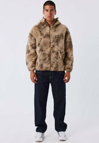 Giovane uomo che indossa una giacca in pile zipata marrone a fantasia, jeans scuri larghi, sneakers bianche, in piedi con le mani rilassate sui fianchi.