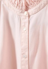 Svetloružová blúzka s gombíkmi na prednej strane, s nariaseným výstrihom a hladkou textúrou. Šesť bielych gombíkov pozdĺž zapínania.