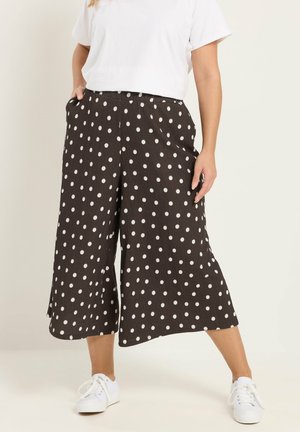 Kvinde iført sorte vidde-ben korte bukser med hvide polkaprikker, hvid t-shirt og hvide sneakers, stående med hænderne i lommerne.