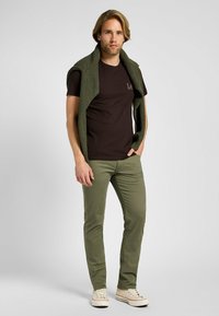 Camiseta marrón con logo, chaleco de punto verde oliva sobre los hombros y pantalones verde claros. Zapatillas beige completan el atuendo.