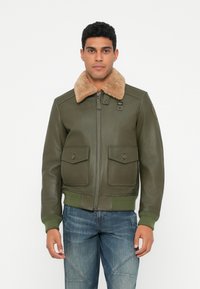 Uomo che indossa un giaccone di pelle verde oliva con colletto in shearling beige e jeans blu, in piedi contro uno sfondo bianco semplice.