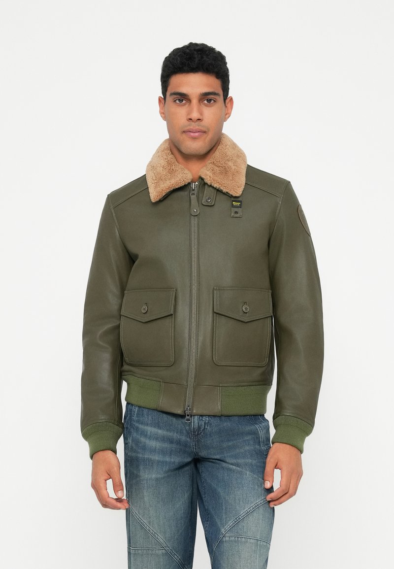 Uomo che indossa un giaccone di pelle verde oliva con colletto in shearling beige e jeans blu, in piedi contro uno sfondo bianco semplice.