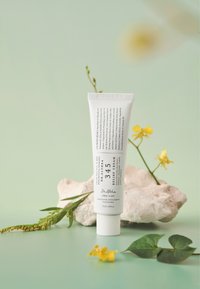 Biała tuba o pojemności 50ml z kremem łagodzącym, z czarnym napisem, położona na teksturowanym kamieniu, otoczona zielonymi liśćmi i żółtymi kwiatami na tle w delikatnym odcieniu zieleni.
