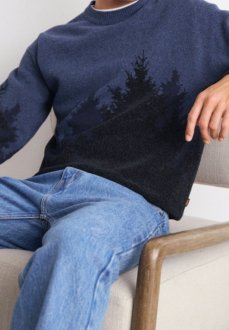 Personne portant un pull bleu foncé avec un motif silhouette d'arbre noir, un jean bleu clair, assise dans un fauteuil beige avec des accoudoirs en bois.