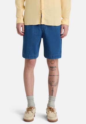 Uomo che indossa una maglietta gialla, pantaloncini di jeans blu, scarpe beige con calzini grigi e tatuaggi alle gambe che includono una faccina sorridente, ali e una spada.