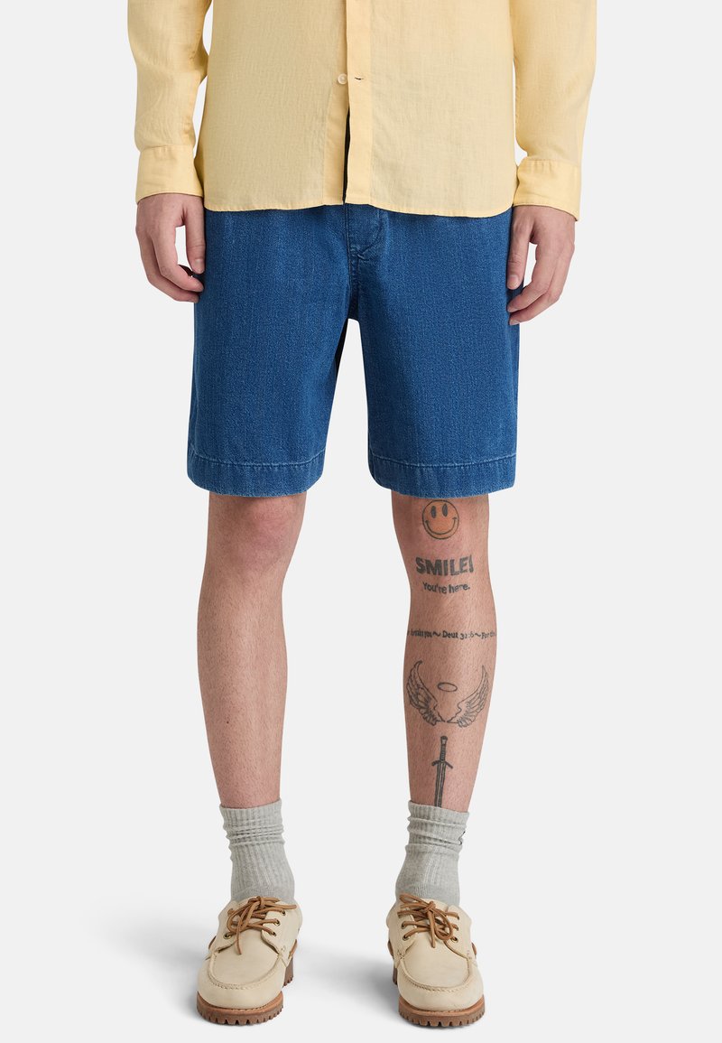 Homme portant une chemise jaune, un short en jean bleu, des chaussures beiges avec des chaussettes grises, et des tatouages sur les jambes comprenant un visage souriant, des ailes et une épée.