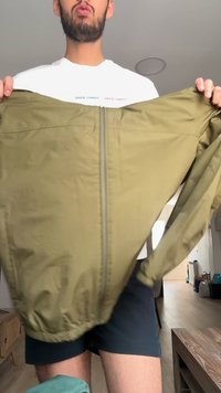 Homme tenant une veste zippée vert olive ouverte tout en se tenant dans une pièce avec un sol en bois et des murs clairs.