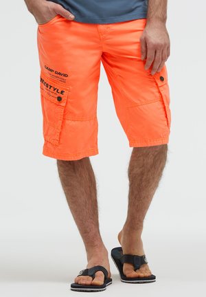 Man wearing bright orange cargo shorts with black text and black flip-flops, standing with one hand in pocket against plain background.
