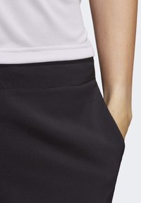 Detailní záběr osoby v bílém topu a černých kalhotách s rukou odpočívaj ící v kapse kalhot.