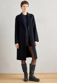 Manteau de marine au design ajusté, avec un col en revers, des boutons sur le devant et une coupe droite, associé à des bottes noires montantes.