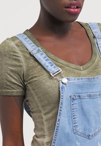 Grünes V-Ausschnitt-T-Shirt, kombiniert mit blauen Jeans-Latzhosen. Die Latzhosen haben verstellbare Träger, eine Vorderasche und eine abgenutzte Optik.