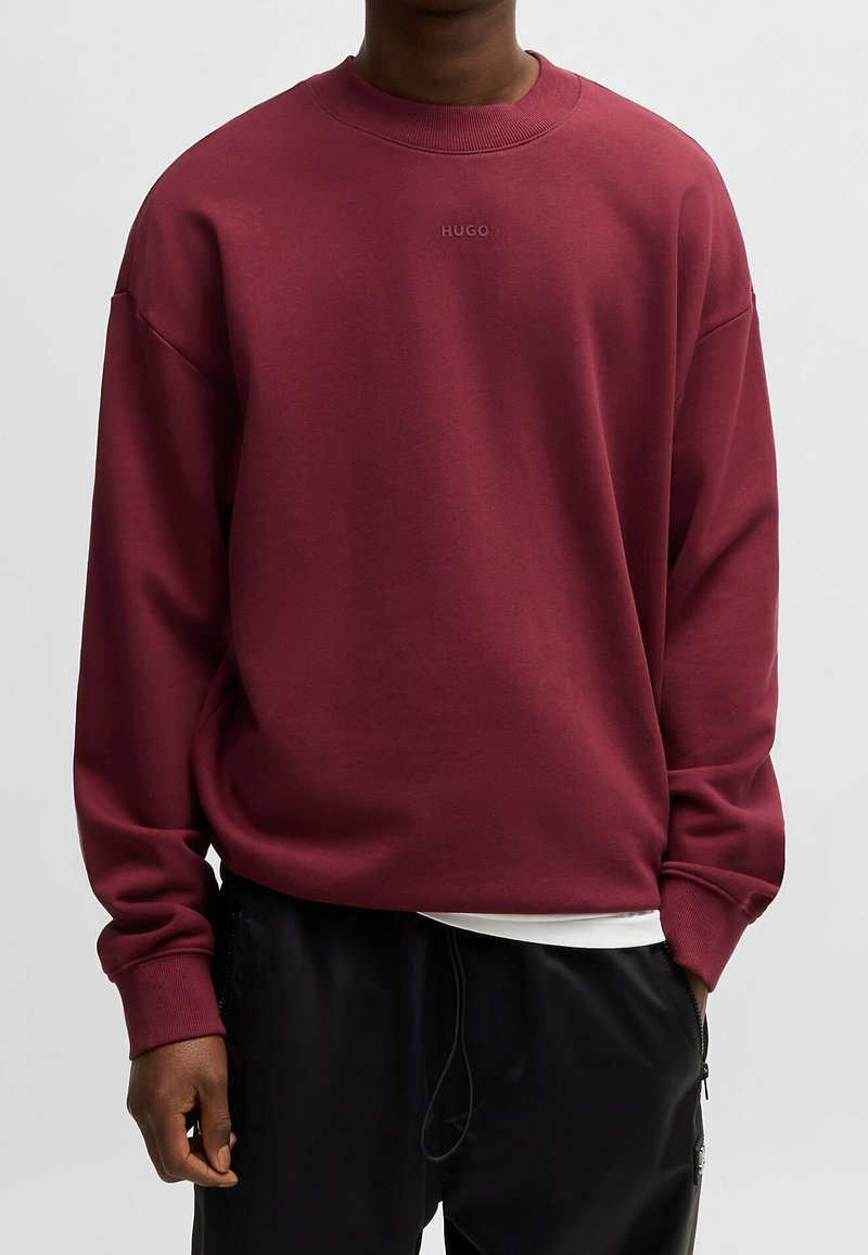 Homme portant un sweat-shirt à manches longues bordeaux avec "HUGO" brodé sur la poitrine et un pantalon noir avec une poche zippée.