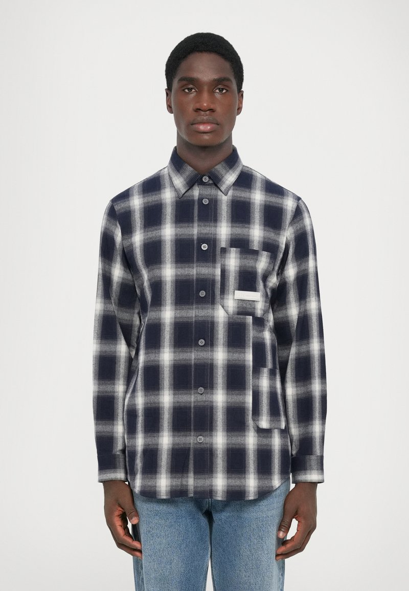 Camisa de flanela xadrez azul-marinho e branca, com fecho de botões na frente, colarinho clássico e um bolso no peito com um detalhe de destaque.