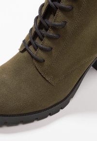 Olijfgroene suède enkellaars met zwarte veters, ronde neus en een gestructureerde rubberen zool. Kenmerkt zich door een gladde afwerking en gestikte details.