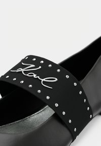 Czarna klapka na płaskiej podeszwie z szerokim paskiem ozdobionym srebrnymi ćwiekami i metalicznym logo "Karl". Gładka, matowa tekstura o nowoczesnym designie.