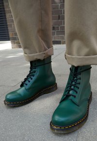 Green leather ankle boots with black laces, yellow stitching, and a chunky black sole. Pants are tan with cuffs rolled up.