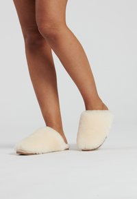Crèmekleurige, fluffy slippers met een zachte textuur, open achterkant en een platte tan kleurige zool, opgesteld op een lichte achtergrond.