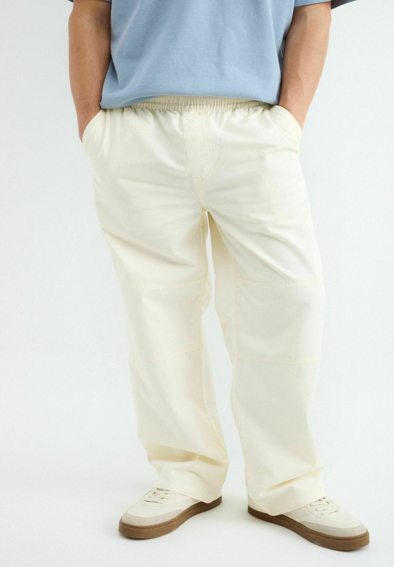 Personne portant un pantalon ample beige clair avec une taille élastique et des baskets blanches à semelles marron, mains dans les poches, sur fond uni.