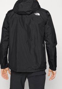 Black hooded jacket with elastic cuffs worn by person, featuring white The North Face logo on upper back right side.