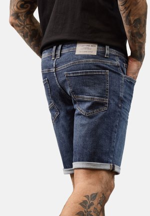 Mann trägt hochgekrempelte blaue Jeansshorts und ein schwarzes Hemd, mit sichtbaren Tätowierungen an Armen und Beinen, steht mit den Händen in den Taschen.