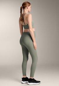 Olivno zeleni športni modrček s križnimi naramnicami in visokim pasom, ujemajoče se legice. Model nosi črne športne čevlje. Gladka tekstura.
