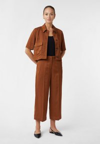 Veste brune courte à manches courtes avec fermeture à bouton et deux poches avant, associée à un pantalon droit taille haute et des ballerines à talon noires.