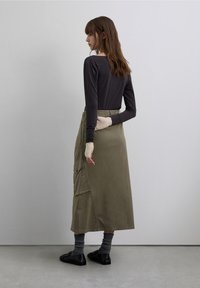 Top de manga larga oscuro combinado con una falda verde oliva hasta la rodilla, con cintura elástica y bolsillos laterales. Se ven zapatos negros y calcetines grises.