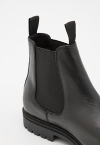 Botín Chelsea de cuero negro con paneles laterales elásticos, lengüetas y una suela resistente. Textura suave con detalles de costura visibles.