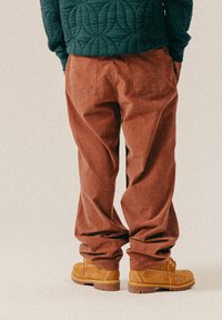 Braun karierte Hosen mit lockerer Passform, strukturiertem Stoff und zwei Gesäßtaschen. Kombiniert mit hellbraunen Stiefeln mit gelben Schnürsenkeln und robusten Sohlen.