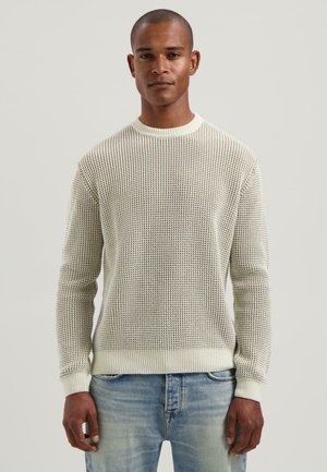 Man in een lichtbeige gewoven trui en vervaagde blauwe spijkerbroek, staande tegen een effen lichte achtergrond.