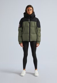 Doudoune en vert olive avec des accents noirs, fermeture éclair, col montant et patch logo sur la manche. Associée à des leggings noirs et des baskets blanches.