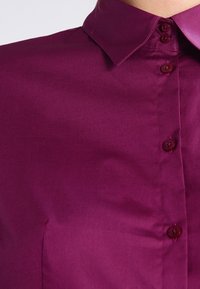 Camicia a maniche corte di un profondo magenta. Tessuto liscio, design classico con bottoni abbinati e colletto a punta.