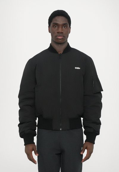 Schwarze Bomberjacke aus wattiertem Material mit gerippten Bündchen und Saum, ausgestattet mit einem Reißverschluss und einer Tasche am linken Ärmel. Markenlogo auf der Brust.