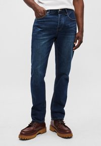 Blugi din denim întunecat, cu un design drept, stil cu cinci buzunare și cusături vizibile. Asortați cu încălțăminte din piele maro cu șireturi.