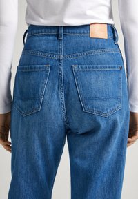 Jeans de mezclilla azul oscuro con cintura alta, que cuentan con dos bolsillos traseros y una etiqueta de cuero color marrón claro en la cinturilla. Textura suave y corte clásico.