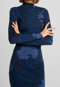 Long-sleeved dress in dark navy with floral pattern in varying shades of blue. High neckline and form-fitting silhouette. Smooth texture.