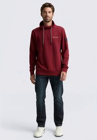 Bordeauxroter Kapuzenpullover mit Kordelzug, Kängurutasche und Logodetail; kombiniert mit dunkler Jeans und weißen Sneakers.