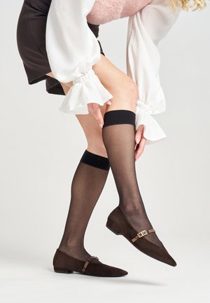 Ballerine in suede marrone con punta affilata e una cinghia decorativa. Modella che indossa calze trasparenti nere fino al ginocchio. Tessuto morbido e testurizzato.