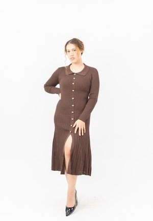 Vrouw poseert in een lange bruine geribde jurk met knopen aan de voorkant en een split, zwart puntige hoge hakken dragend.