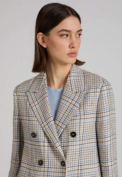 Jonge vrouw met kort bruin haar, gekleed in een beige geruite double-breasted blazer over een lichtblauw shirt, kijkt opzij.