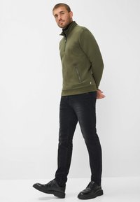 Pullover in pile verde oliva con colletto alto, tasca zippata frontale e polsini a costine, abbinato a jeans neri slim-fit e scarpe nere.