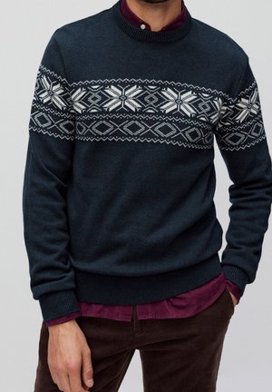 Mann trägt dunkelblauen Pullover mit weißem geometrischem Nordisch-Muster, burgunderfarbenes Hemd mit Knöpfen und braune Hose, Hände in den Taschen.