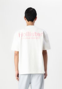 T-shirt blanc à manches courtes avec un grand logo rose "Hollister" et le texte "Designs Originaux" à l'arrière, fabriqué en coton doux.