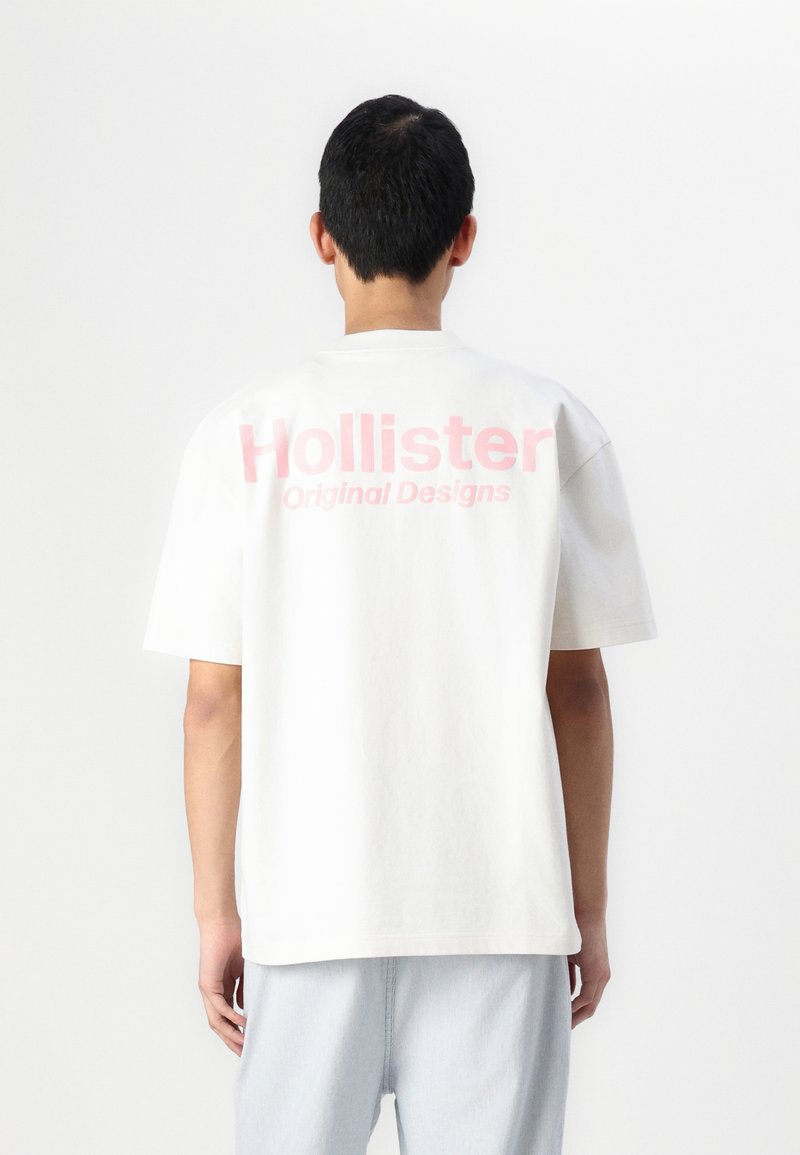 T-shirt blanc à manches courtes avec un grand logo rose "Hollister" et le texte "Designs Originaux" à l'arrière, fabriqué en coton doux.