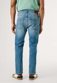 Vaqueros de mezclilla azul claro con corte recto, bolsillos traseros y un parche de cuero marrón en la cintura, combinados con zapatos blancos.