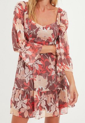 Vrouw die een bloemenjurk draagt met lange mouwen, een vierkante halslijn, pofmouwen en een gerimpelde zoom in tinten rood, roze en beige.