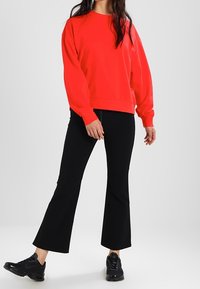 Sudadera de forro polar roja con cuello redondo y hombros caídos, combinada con pantalones negros acampanados y zapatillas negras. Textura suave.